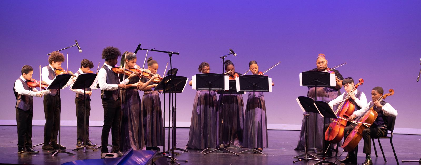 A youth string ensemble of violinists and cellists performs at music stands before a purple backdrop.